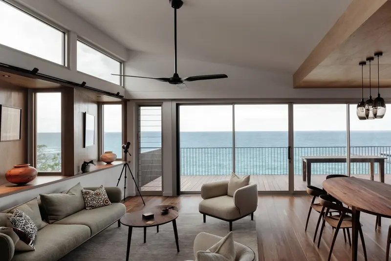 Open-plan living and dining room with timber floors and a raked ceiling, opening through glass doors to a deck above the ocean